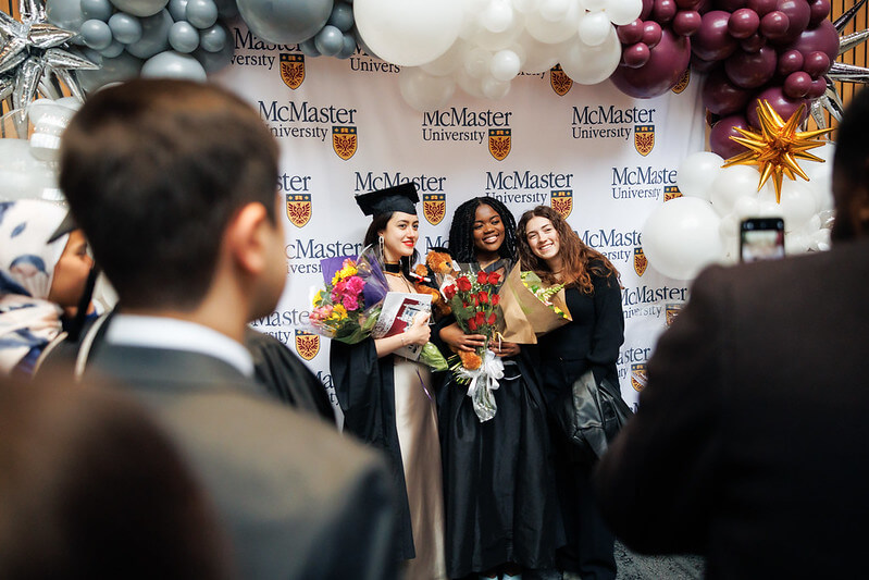 Three graduates in regalia stand in front of a McMaster branded backdrop. They are holding flowers and surrounded by a balloon arch.