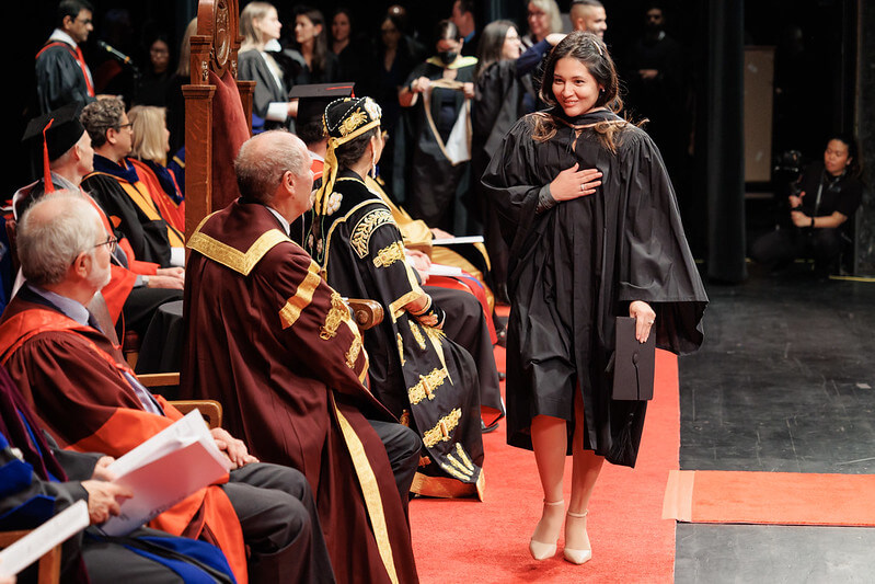 A graduate in a graduation robe and hood is walking across the stage in front of the Chancellor's Party. They are holding their hand over their heart to express their gratitude.