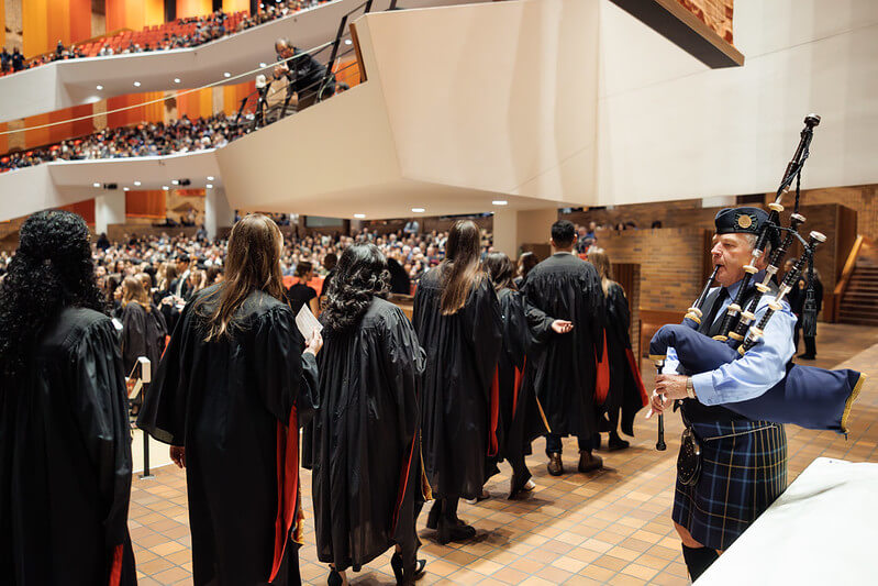 A line of graduates in graduation regalia are shown lining up from the back, heading into an auditorium hall where guests stand in their seats. A bagpiper is to the right of the image in traditional wear.